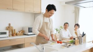 実家暮らしの38歳日本人男性が、エプロンを着用し、台所で野菜を切るなど自律して夕食を調理している様子。親は見守るだけで、男性が主体的に家事をこなしている。